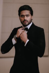 Portrait of a stylish man in a black tuxedo adjusting his bow tie indoors.