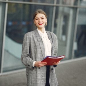 Positive young woman in trendy wear smiling at camera while holding documents standing against glass building in downtown